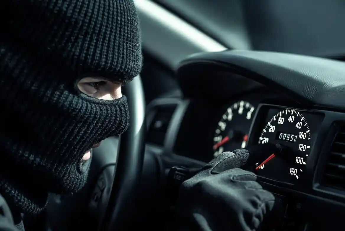 A person wearing a black balaclava and gloves leaning into a car and tampering with the odometer, which displays a falsified low mileage reading — illustrating odometer fraud