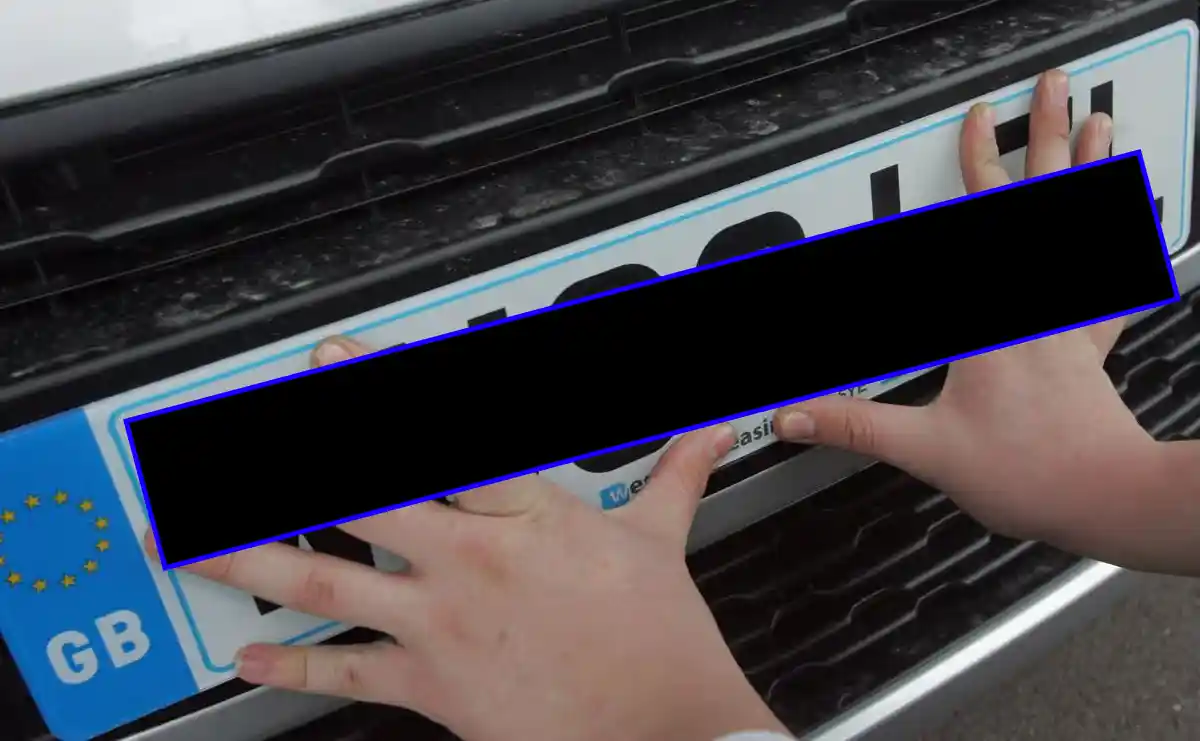 Close-up of two hands fitting a UK number plate with a GB Euro band to the front of a car, with the registration number deliberately concealed behind a solid black bar — illustrating a listing photo where the plate has been hidden so it cannot be checked