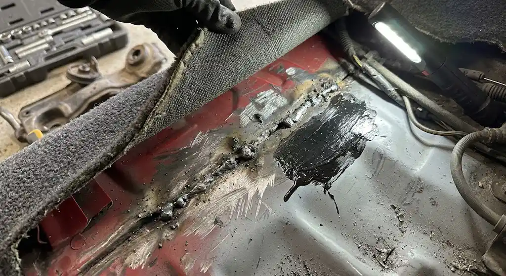 Close-up under a car's interior carpet showing a rough, non-factory weld seam running across the floor pan with grinder marks and fresh underseal — the classic telltale of a cut and shut joint